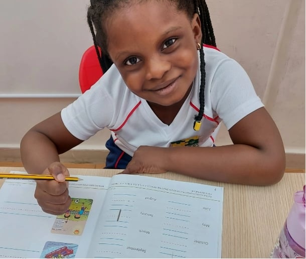 A girl from Bright Learners - American School in Dubai smiles at the camera while doing homework